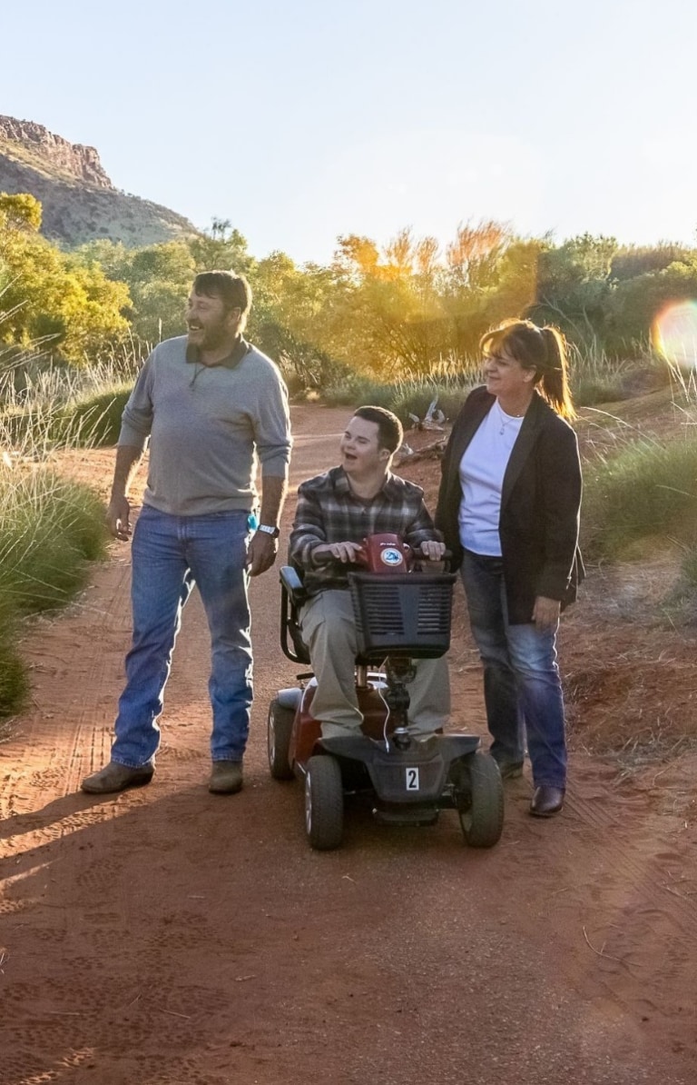 Man seated on a mobility device with another man and woman, wandering among the red dirt and greenery of Alice Springs Desert Park with the sun in the background in Alice Springs, Northern Territory © Tourism NT/Helen Orr